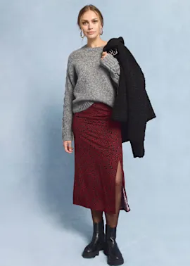 Grauer Pullover, roter Rock mit Muster, schwarzer Mantel und klobige Stiefel.