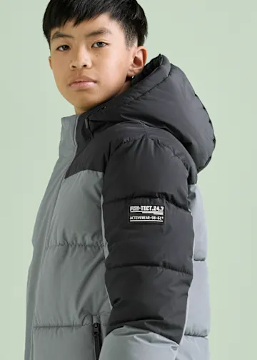 Der Junge trägt eine graue und schwarze Steppjacke mit Kapuze.