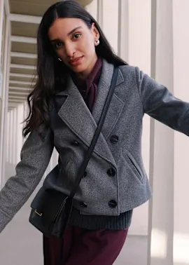 Sie trägt einen eleganten grauen Kurzmantel über einem weinroten Oberteil, dazu eine schwarze Tasche.