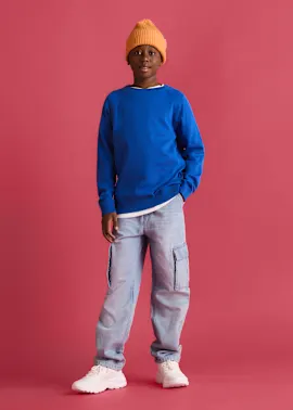 Der Junge trägt eine orange Strickmütze, blaues Sweatshirt, helle Cargohose und weiße Sneaker.