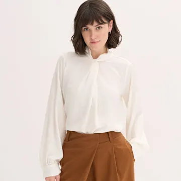 Weiße Bluse mit Schleifenkragen, kombiniert mit einer braunen Hose.