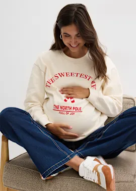 Schwangere Frau in weißem Weihnachtspullover hält liebevoll ihren Babybauch und lächelt glücklich.