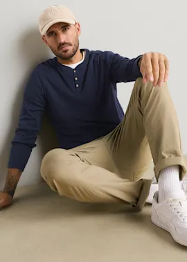 Beige Mütze, blaues Henley-Shirt, beigefarbene Hose und weiße Sneaker sind zu sehen.