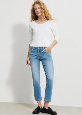 Sie trägt ein weißes Langarmshirt, eine hellblaue Jeans und schwarze Loafer.