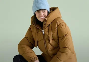 Sie trägt eine hellblaue Mütze und eine stylische braune Steppjacke mit Kapuze.