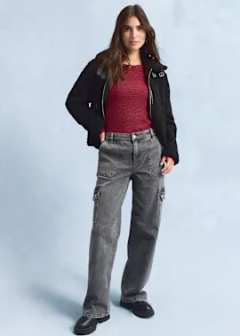 Schwarze Jacke, rotes Oberteil, weite graue Jeans und dunkle Schuhe.