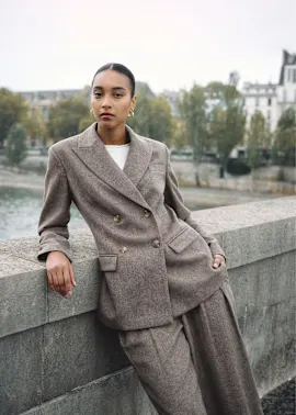 Eine Frau in einem eleganten braunen Anzug sitzt an einem Flussufer in Paris.