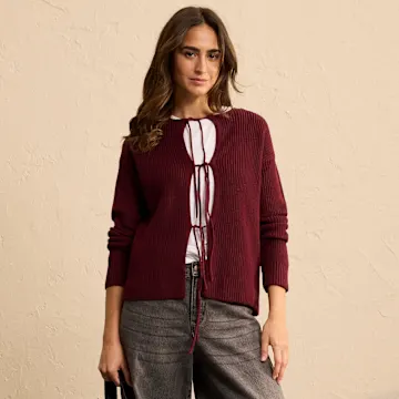 Weinrote Strickjacke über weißer Bluse mit Bändchen. Graue Hose für eleganten Kontrast.