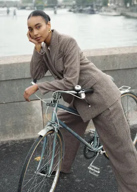 Elegante Frau in beigem Mantel sitzt nachdenklich mit ihrem Fahrrad am Flussufer.