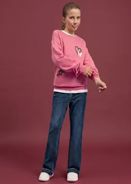 Sie trägt ein pinkes Sweatshirt mit Herzmotiv, blaue Jeans und weiße Sneaker.