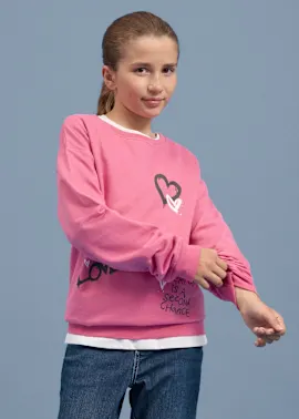 Mädchen trägt ein pinkes Sweatshirt mit Herz-Motiv, dazu Jeans und weißes Shirt.