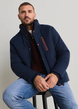 Dunkelblaue Steppjacke mit orangefarbenen Details, roter Strickpullover und helle Jeans.