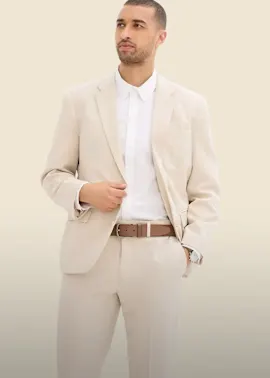 Er trägt einen eleganten beigen Anzug, ein weißes Hemd und einen braunen Gürtel.