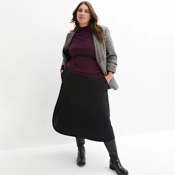 Grauer Blazer, lila Pullover und langer schwarzer Rock mit schwarzen Stiefeln.