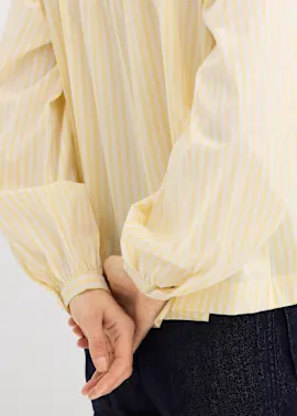 Die gelb-weiß gestreifte Bluse mit bauschigen Ärmeln passt zur dunklen Hose.