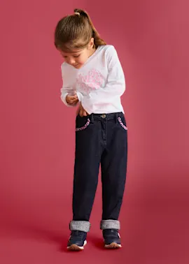 Weißes Herz-Shirt, dunkle Jeans mit gemusterten Taschen und umgeschlagenem Saum, dazu Sneakers.