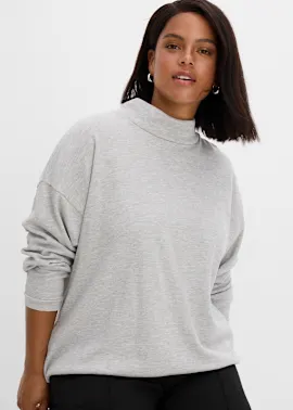 Sie trägt ein bequemes hellgraues Sweatshirt mit Stehkragen und schwarze Hose.