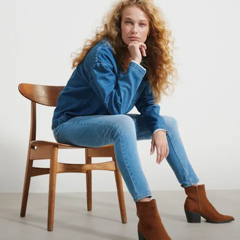 Sie trägt eine blaue Jeansjacke, Jeans und stylische braune Stiefeletten.