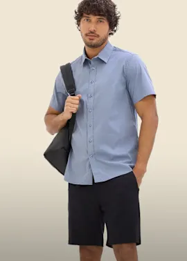 Der Mann trägt ein hellblaues Kurzarmhemd, schwarze Shorts und eine Umhängetasche.