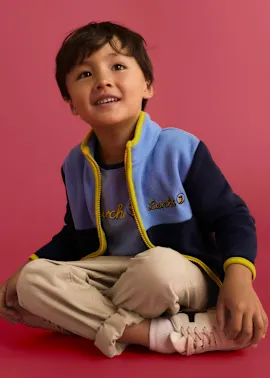 Der Junge trägt eine bunte Fleecejacke, ein hellblaues Shirt, beige Hose und helle Turnschuhe.