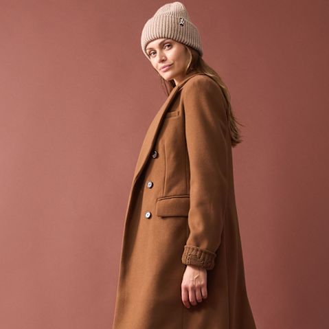 Langer kamelbrauner Mantel, beige Strickmütze und schlichtes Outfit darunter.