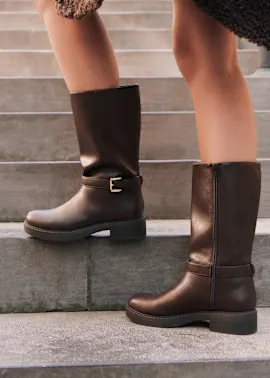 Dunkelbraune Stiefel mit goldener Schnalle und Profilsohle auf steinernen Stufen.