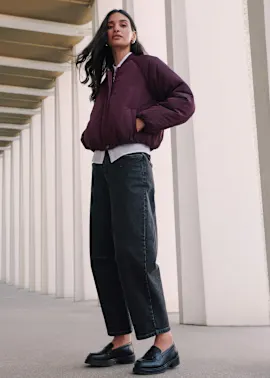Dunkle Kappe, gesteppte bordeauxrote Jacke, roter Cardigan und dunkle Jeans.