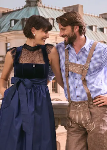 Sie trägt ein elegantes, dunkelblaues Dirndl, er klassische Lederhosen und ein kariertes Hemd.