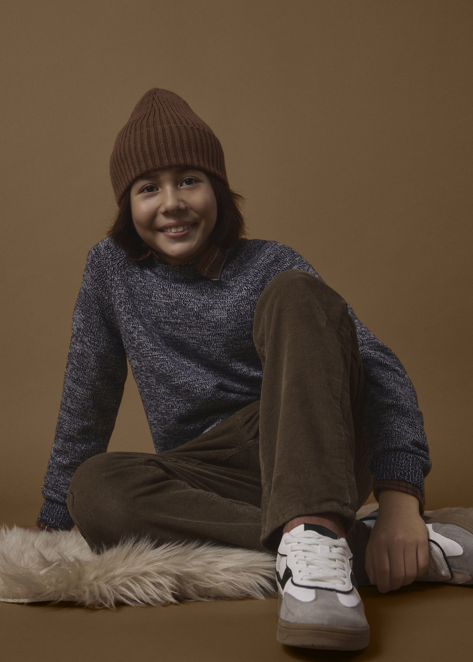 Warm gekleidet mit brauner Mütze, gemustertem Pullover, Cordhose und modernen Sneakern.