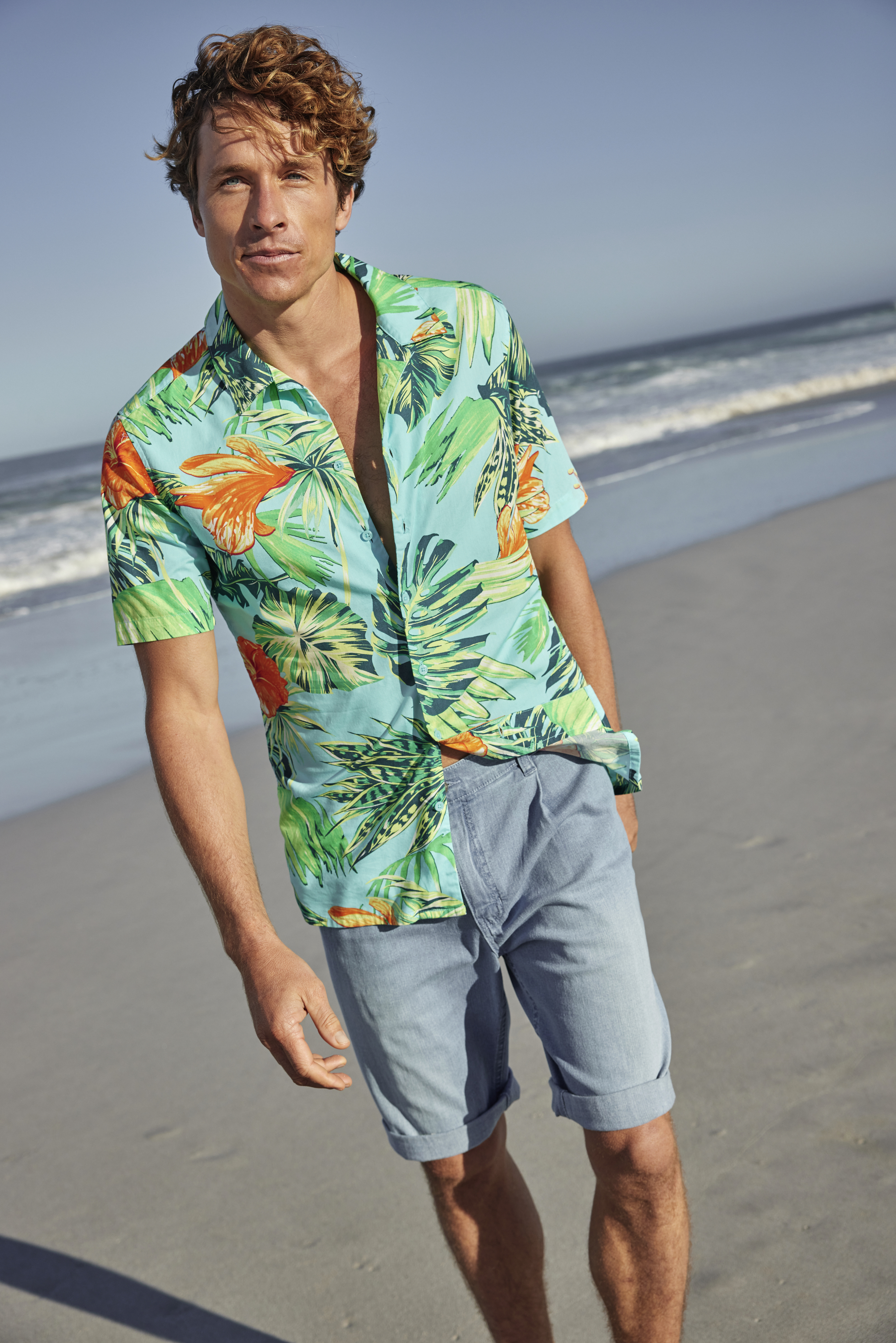 Ein Mann trägt ein buntes Hawaiihemd und helle Shorts am Strand.