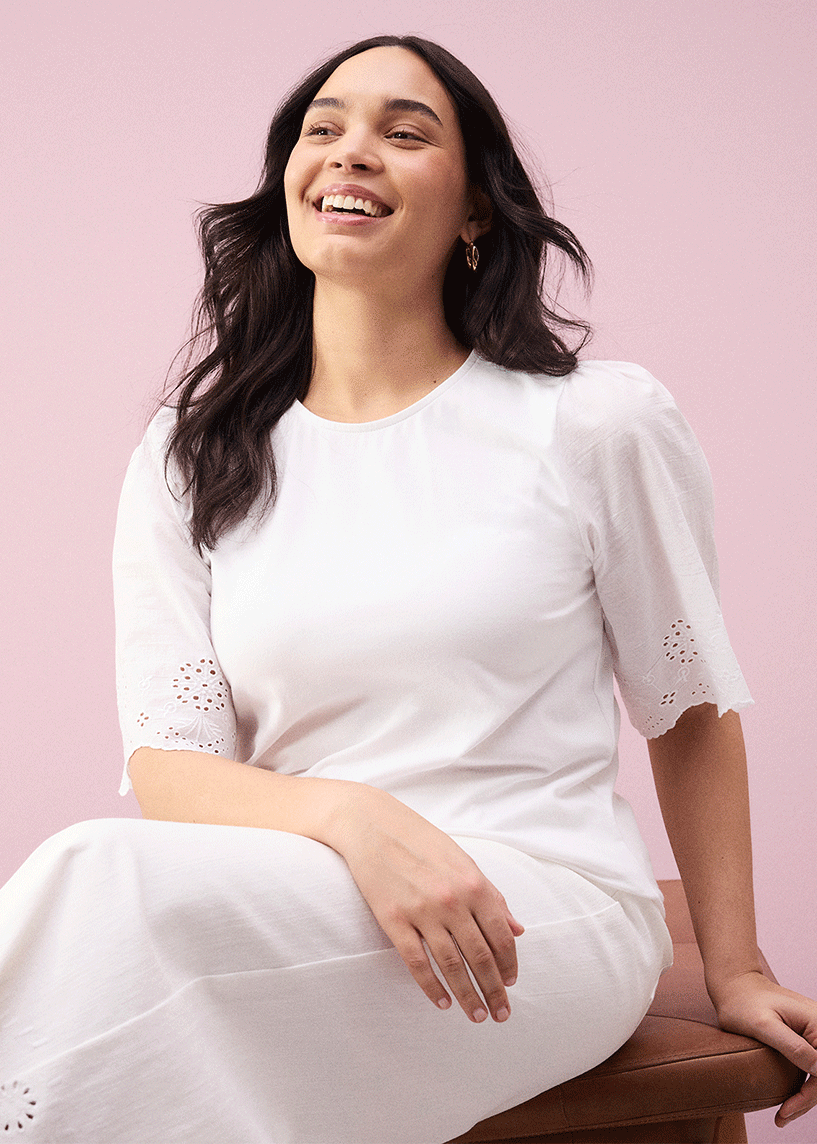 Eine strahlende Frau in einem schlichten weißen Kleid mit kurzen Ärmeln und Lochstickerei.