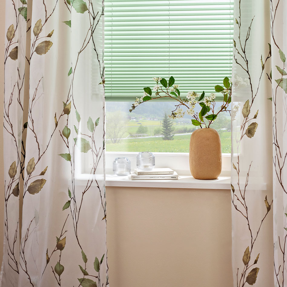 Vorhänge mit Blumenmuster schmücken das Fenster, daneben steht eine schlichte Vase.