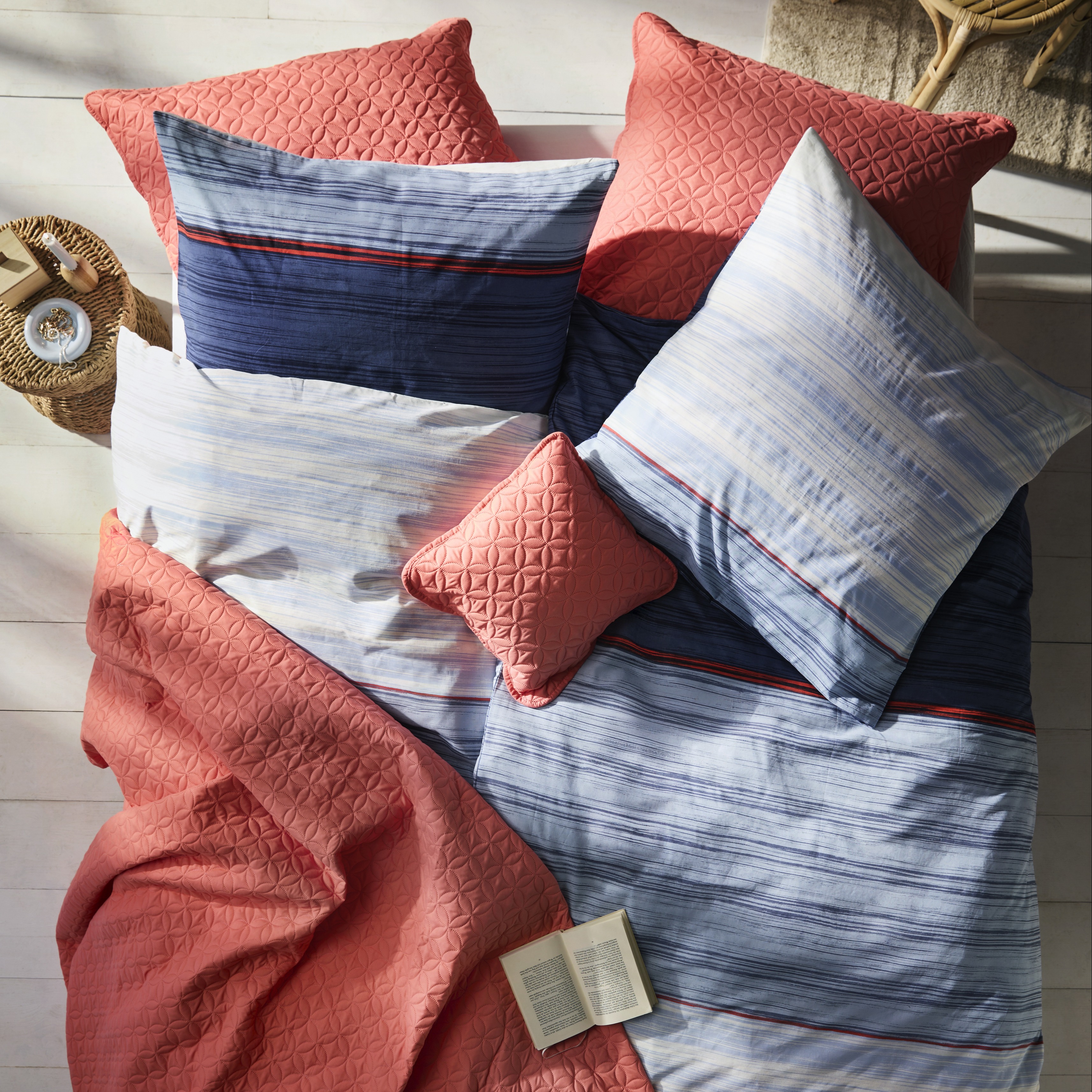 Gestreifte Bettwäsche in Blau und Weiß, kombiniert mit korallenroten Kissen und einer Decke.