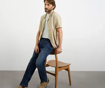Beiges Hemd, graues T-Shirt, blaue Jeans und braune Wildlederschuhe bilden den Look.