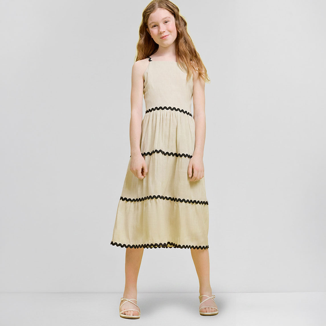 Mädchen in beigem Stufenkleid mit schwarzen Zierbändern und hellen Sandalen.