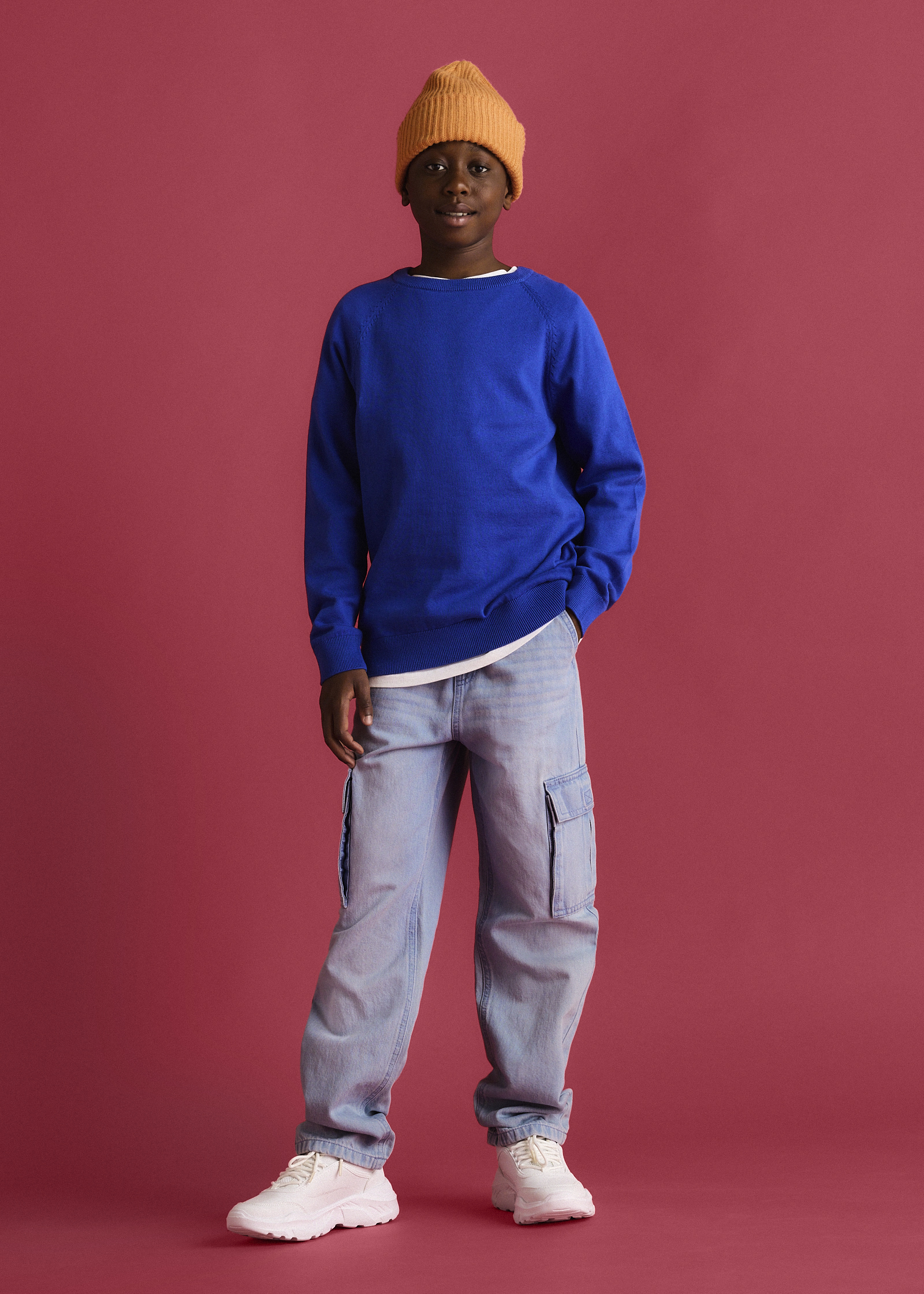 Der Junge trägt eine orange Strickmütze, blaues Sweatshirt, helle Cargohose und weiße Sneaker.