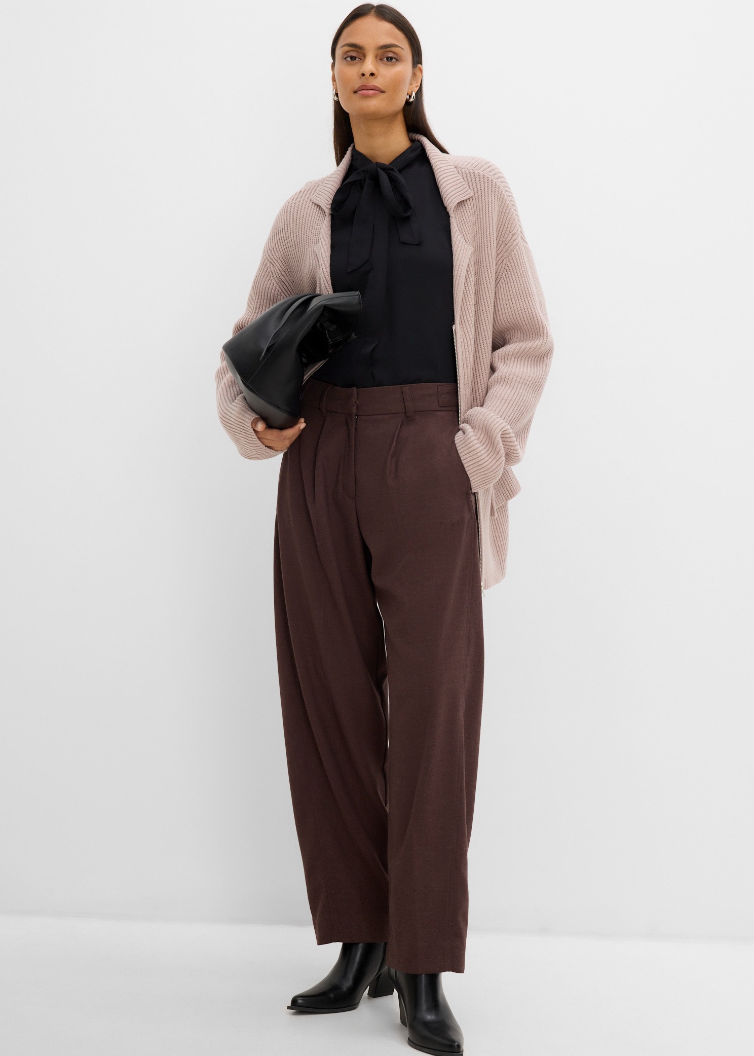 Sie trägt eine schwarze Bluse, beigen Strickcardigan, braune Hose und schwarze Stiefeletten.