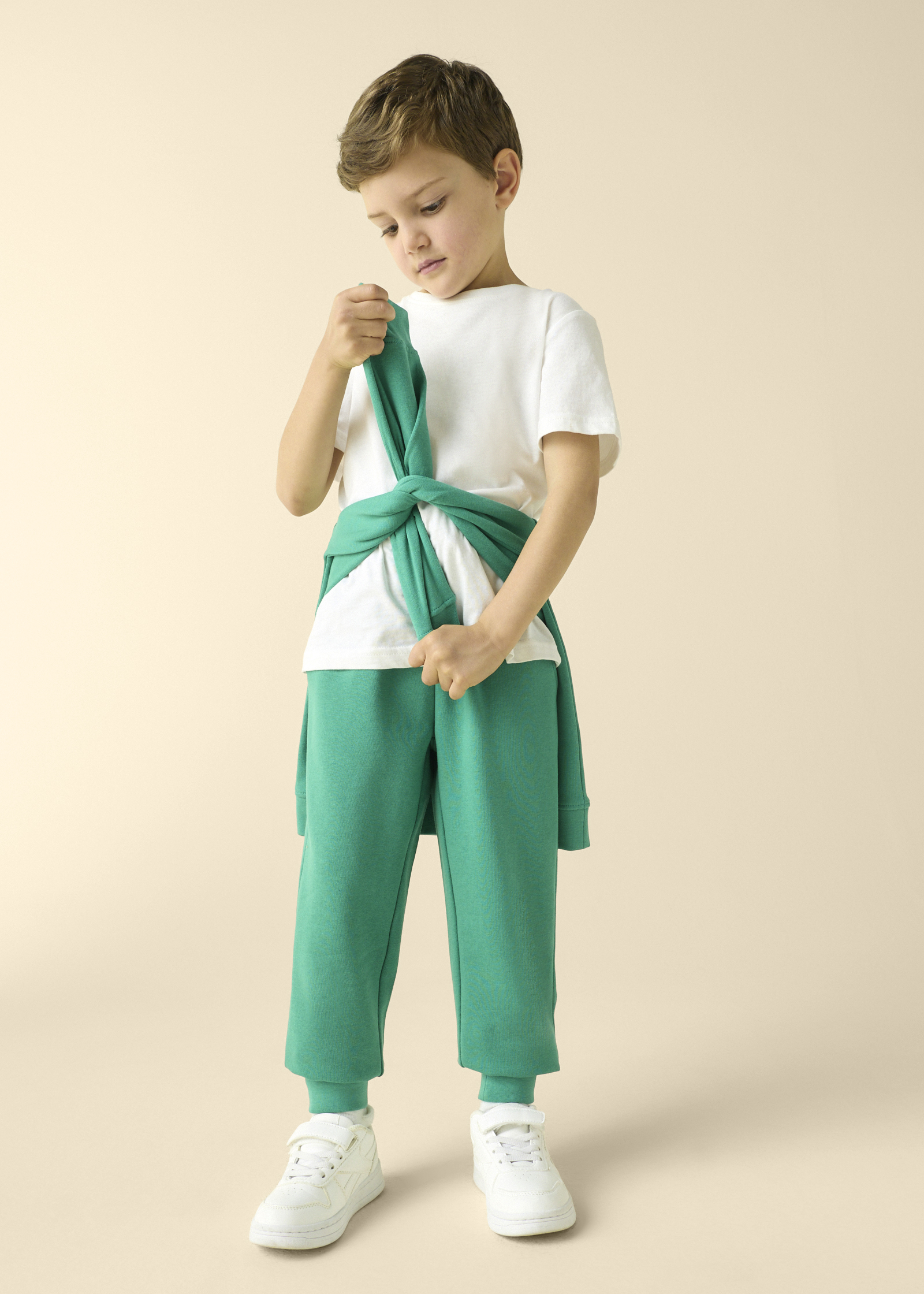 Kind in weißem T-Shirt, grüner Hose und umgebundenem Pullover.
