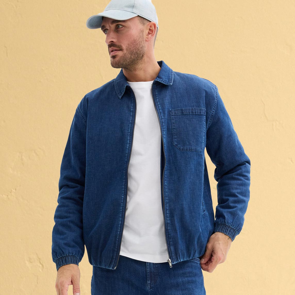 Lässiger Denim-Look mit blauer Jeansjacke, weißem T-Shirt und hellgrauer Baseballkappe.