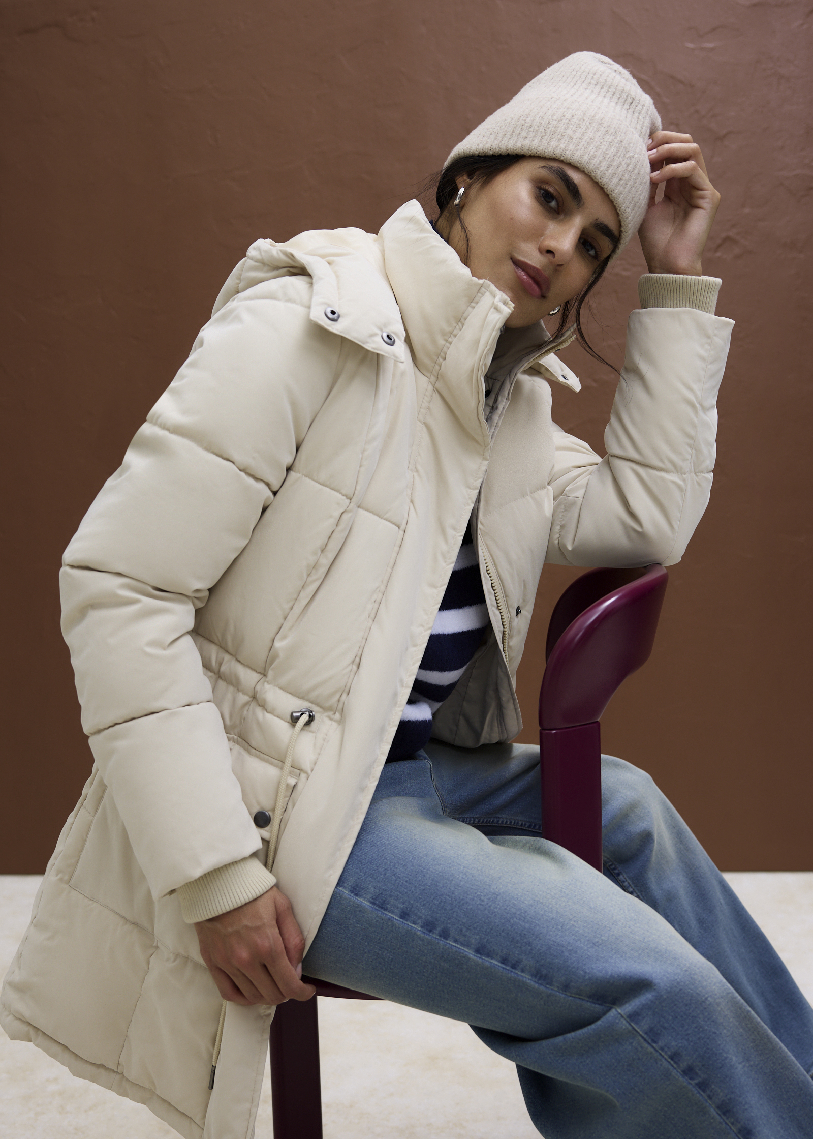 Winterliches Outfit: Cremefarbene Steppjacke, Beanie, gestreiftes Shirt und blaue Jeans.