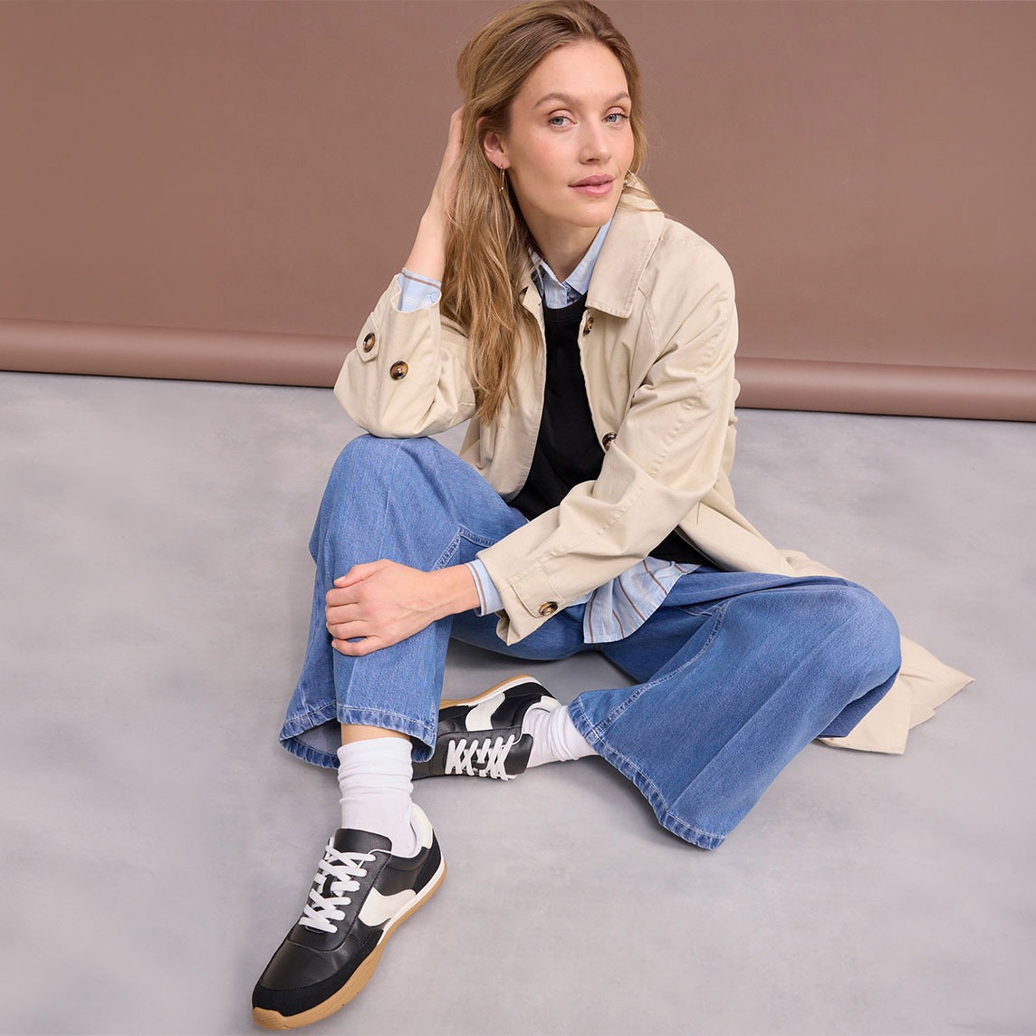 Sitzende Frau mit Trenchcoat, Jeans und schwarzen Sneakern.