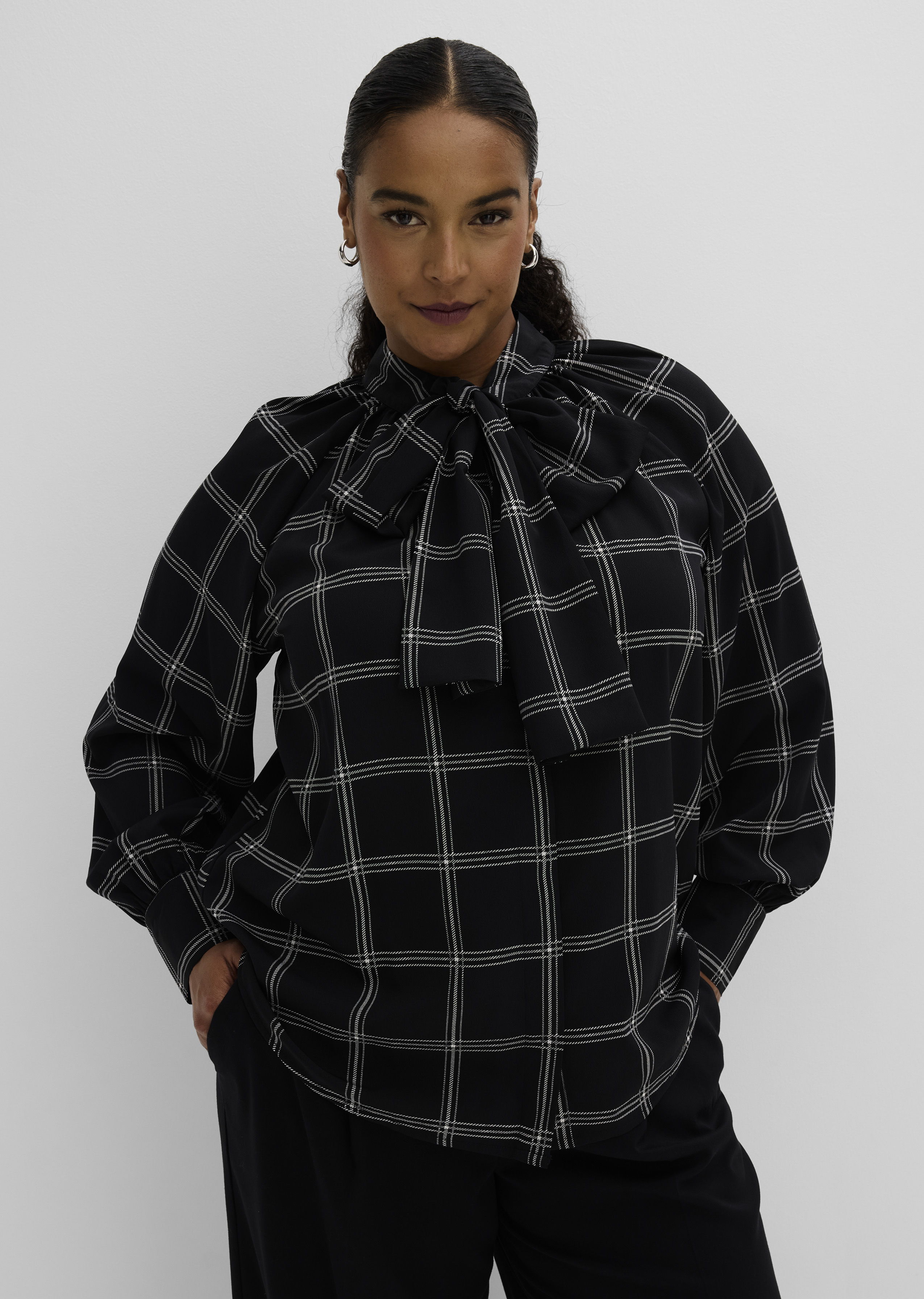 Elegante schwarze Bluse mit weißem Karomuster und großer Halsschleife, dazu schwarze Hose.
