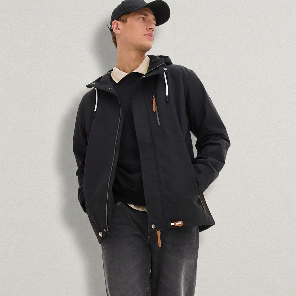 Schwarze Kapuzenjacke, Jeans, Pullover und Cap. 