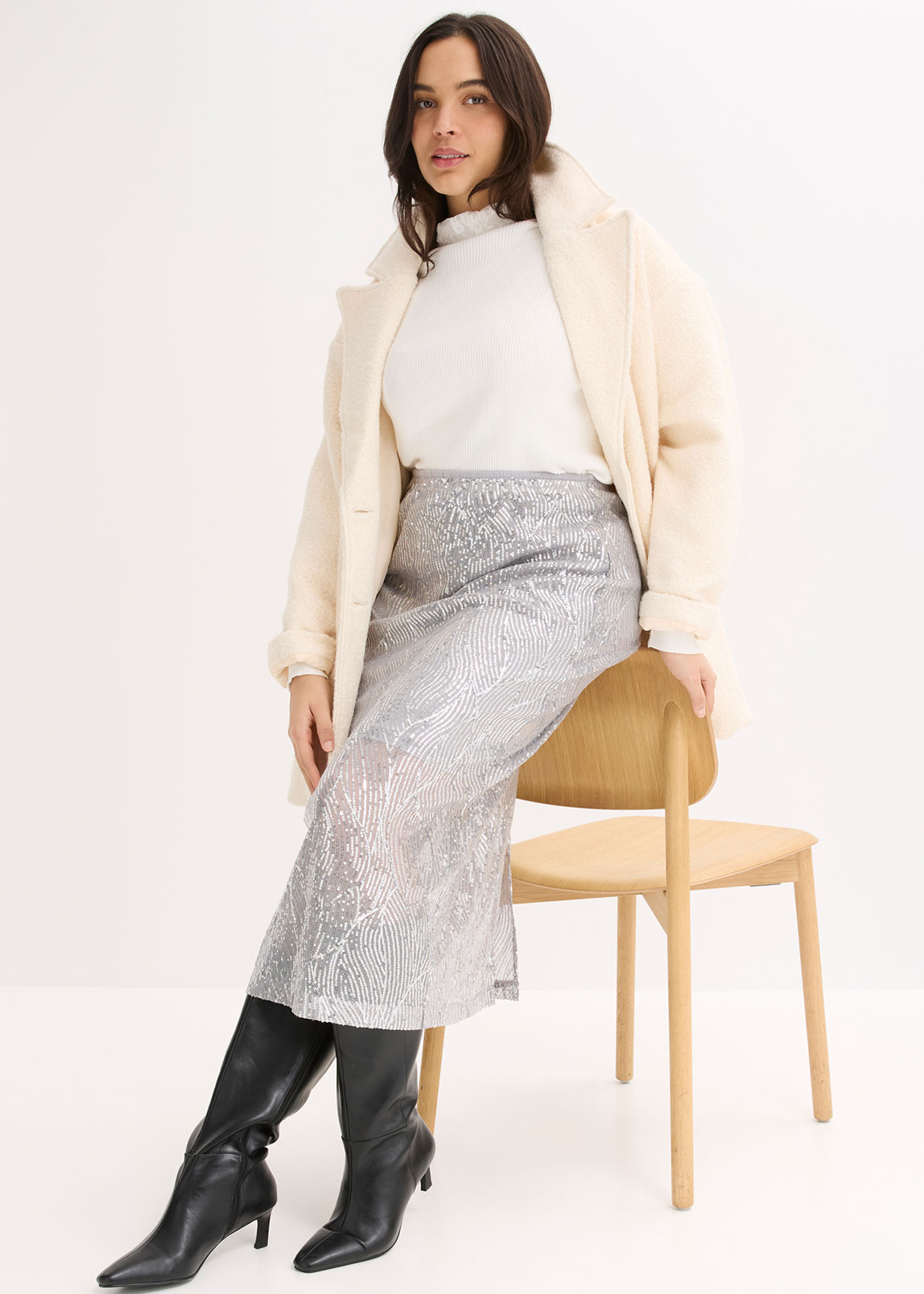 Cremefarbener Mantel, weißer Rollkragenpullover, silberner Rock und schwarze Lederstiefel.