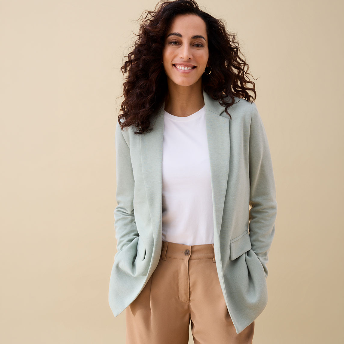 Sie trägt einen mintgrünen Blazer, ein weißes Shirt und eine braune Hose.