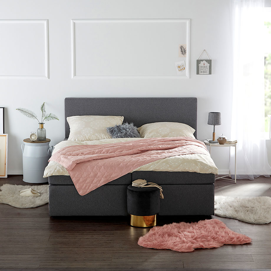 Gemütliches Schlafzimmer mit grauem Bett, rosa Decke und flauschigem Teppich. Elegante Bettwäsche in Weiß.