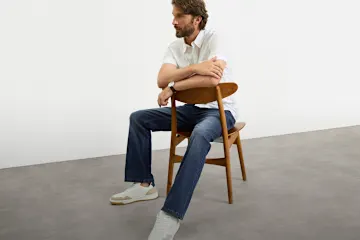 Er trägt ein weißes Hemd, klassische Jeans und stylische helle Sneaker.