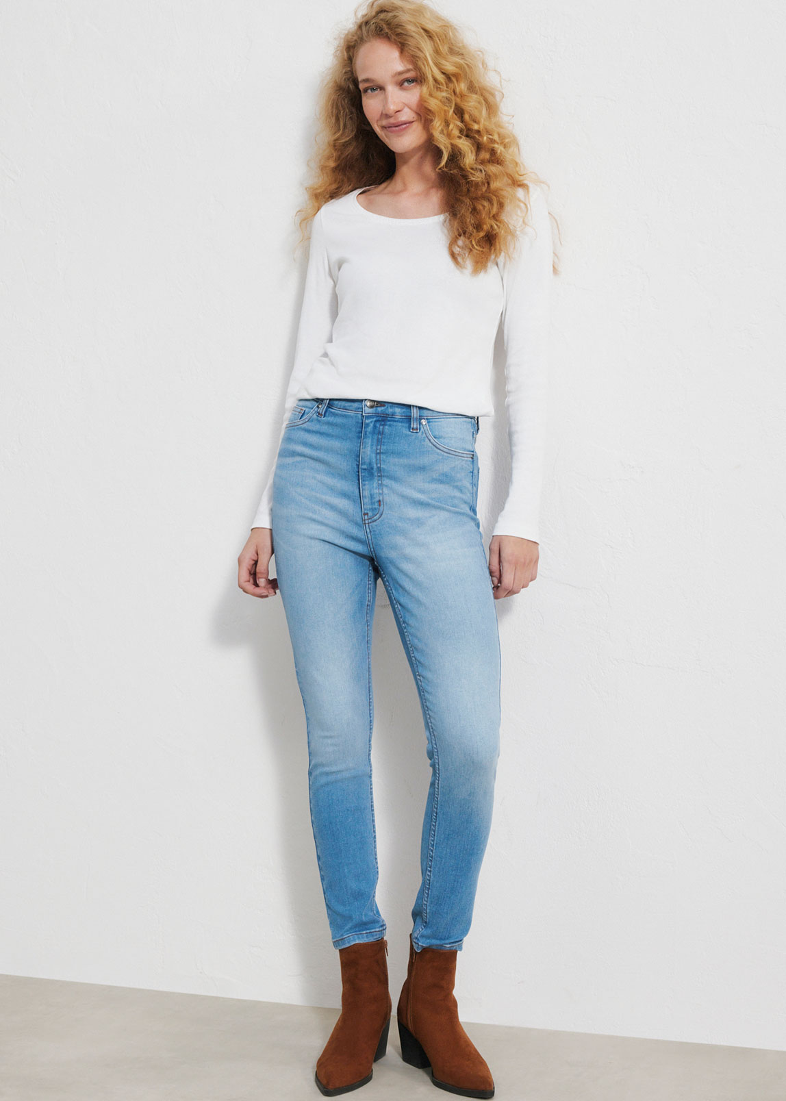 Sie trägt ein schlichtes weißes Oberteil, helle Jeans und braune Stiefeletten.