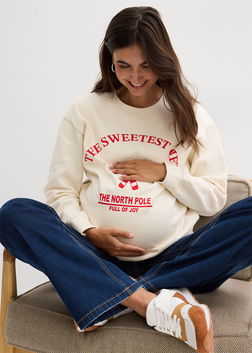 Schwangere Frau in weißem Weihnachtspullover hält liebevoll ihren Babybauch und lächelt glücklich.
