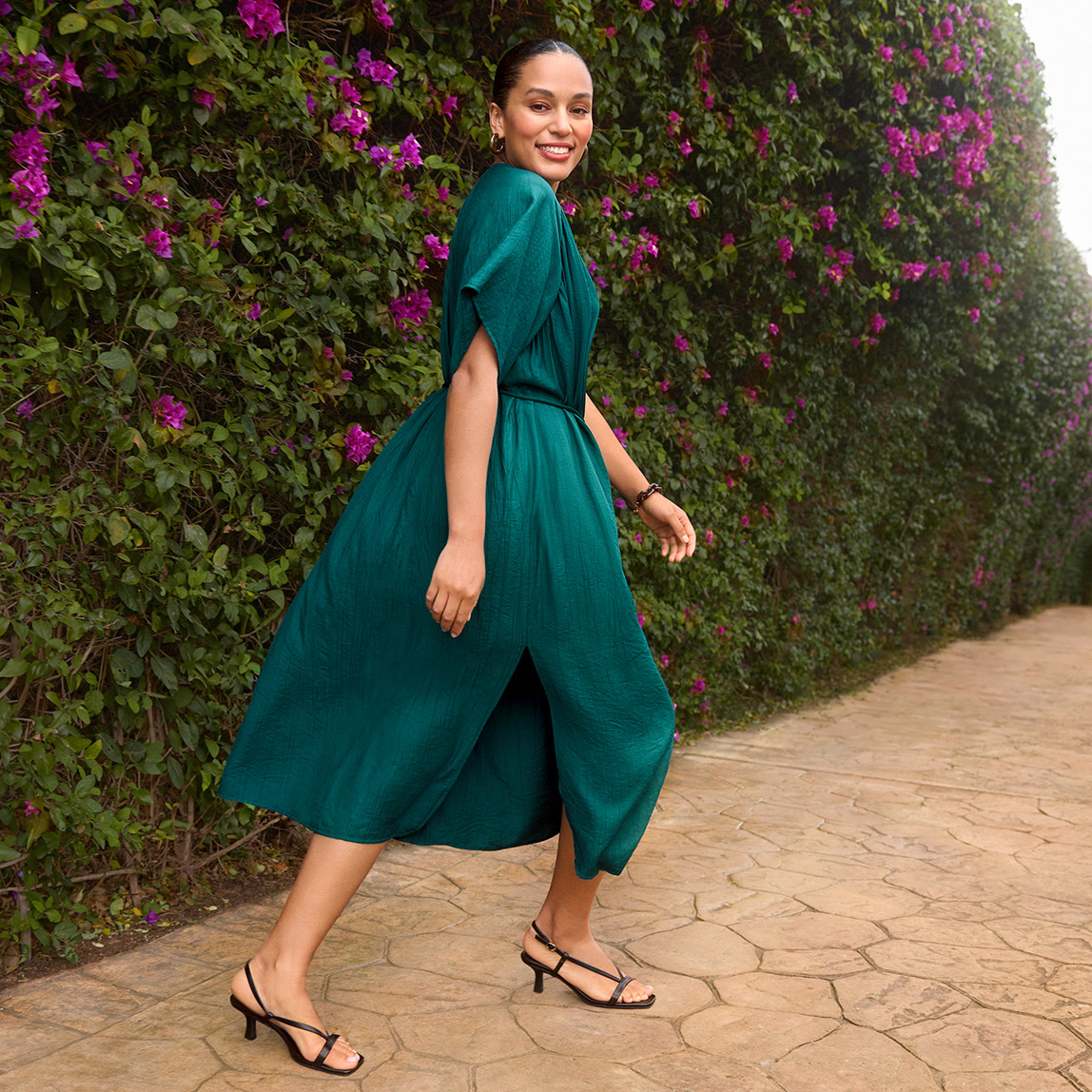 Elegantes türkises Midikleid mit weiten Ärmeln und schwarze Riemchensandalen vor blühender Hecke.